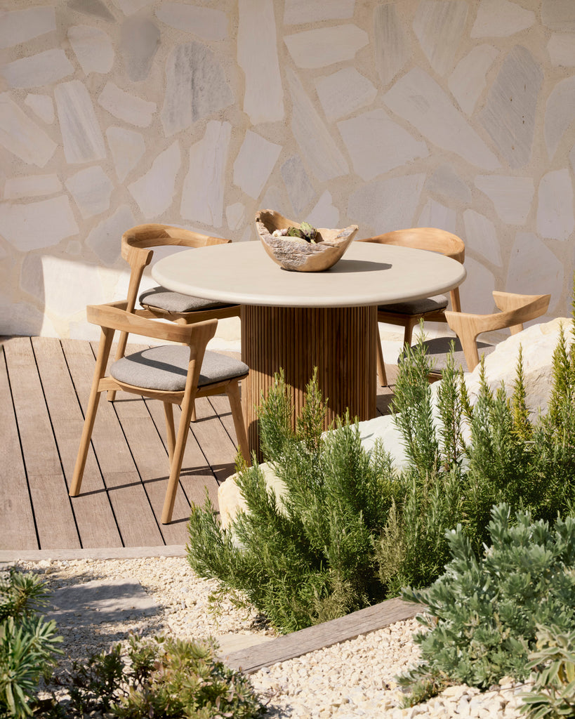 Round outdoor dining table with chairs on a wooden deck against a stone wall.