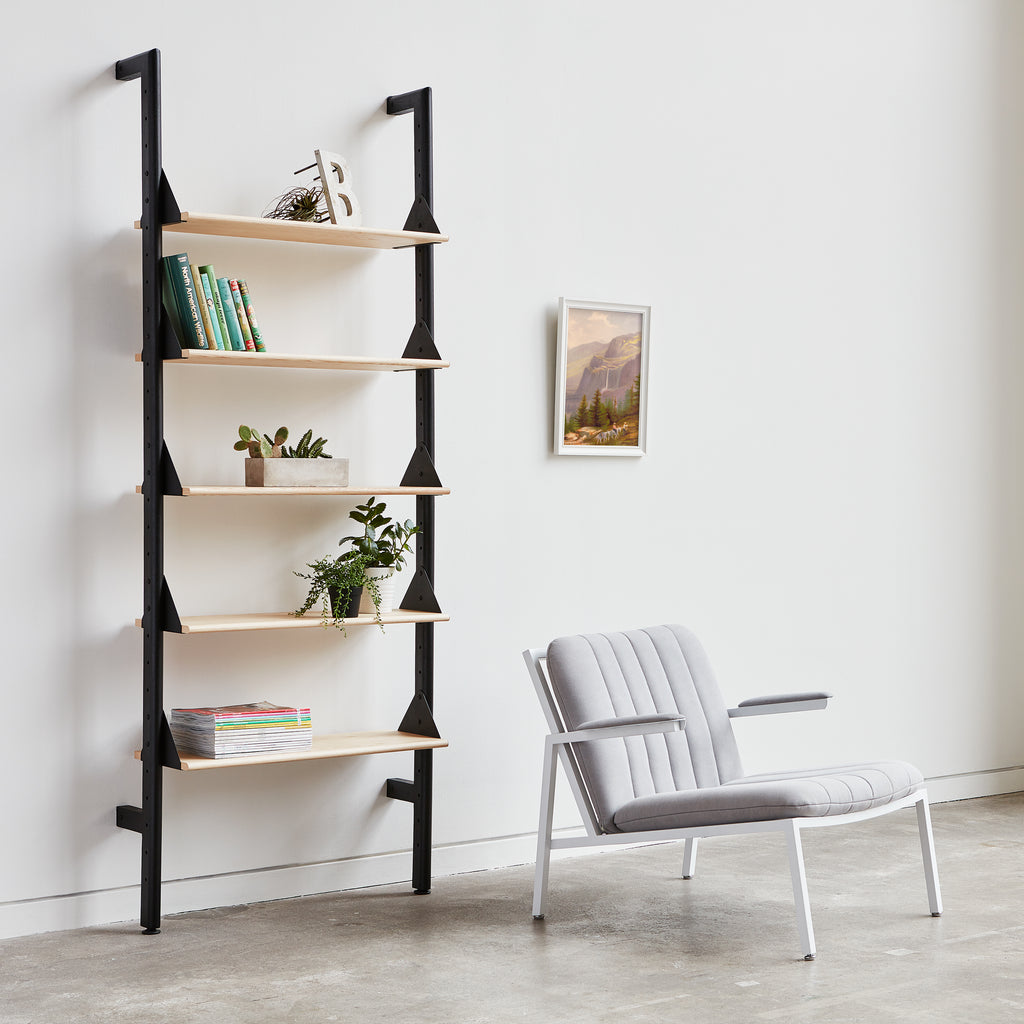 Ladder-style bookshelf with books and plants next to a gray chair in a room.