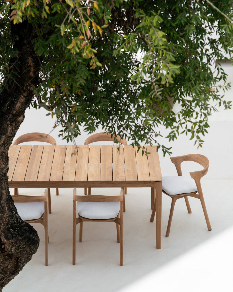 Wooden outdoor dining table with chairs under a tree on a white surface