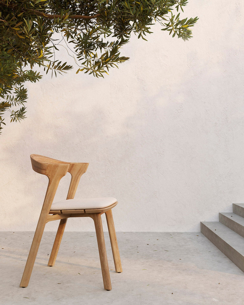 Wooden chair with a white cushion on a light wooden floor with a white wall and greenery in the background.