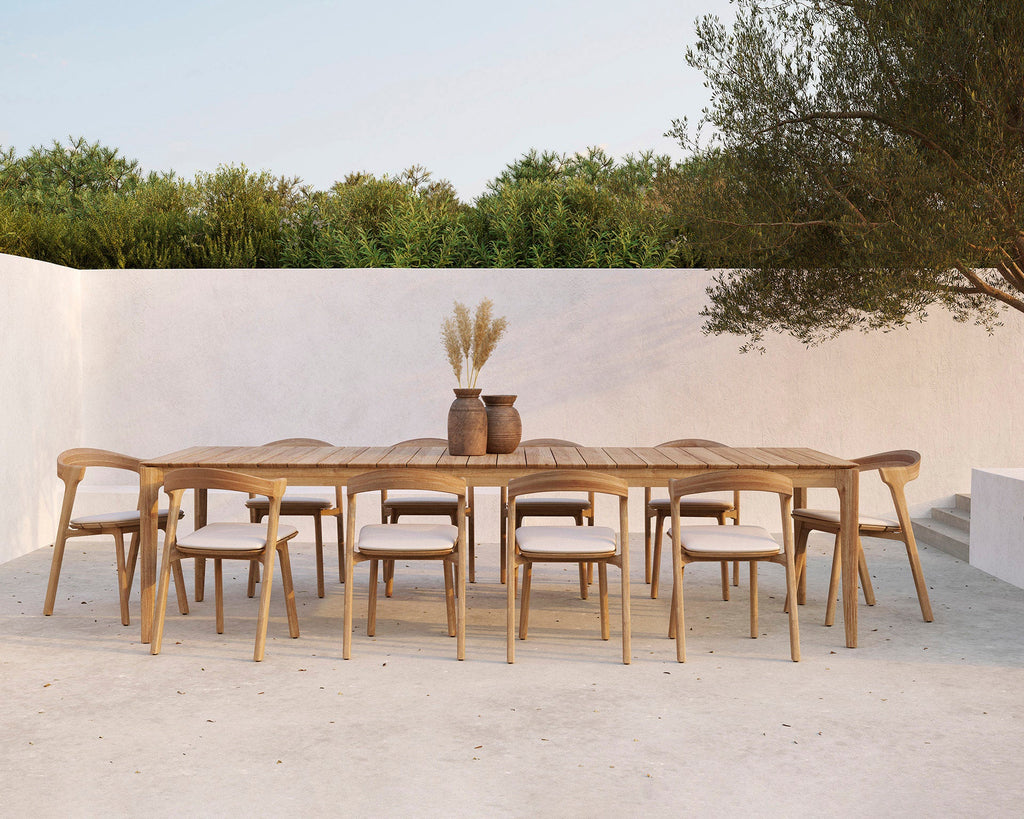 Wooden outdoor dining table with chairs in a serene setting with trees and a white wall.