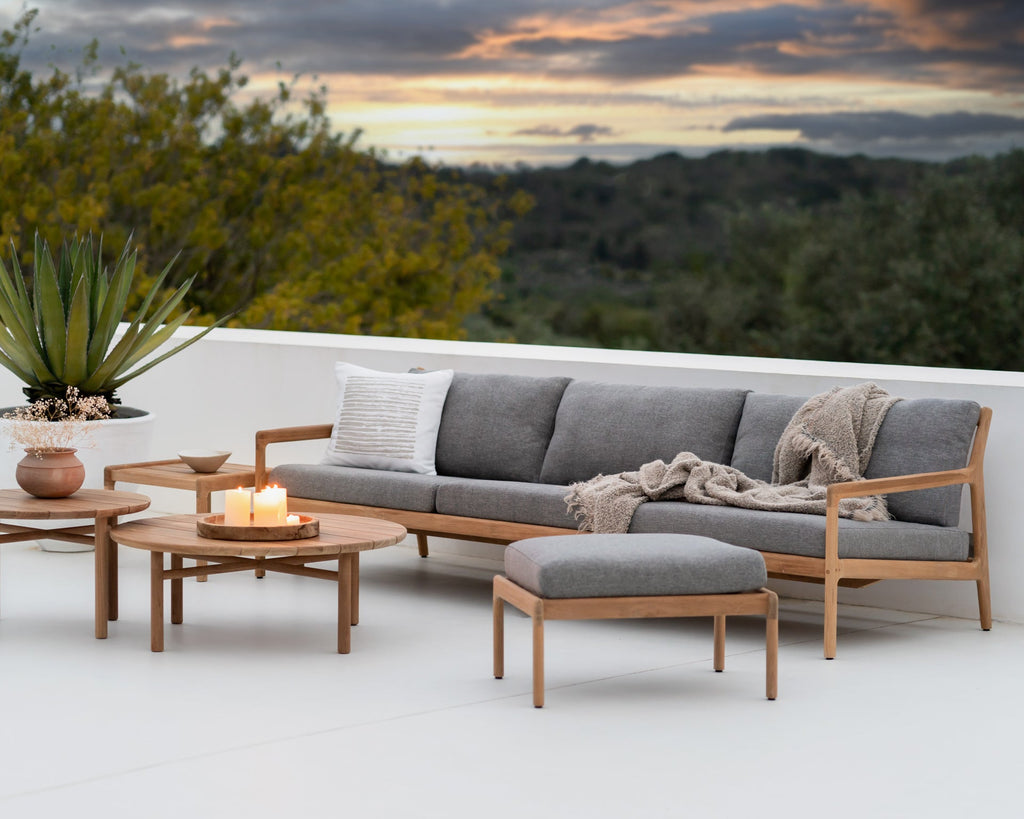 Outdoor patio set with gray sofa, wooden tables, and ottoman on a white deck with a scenic background.